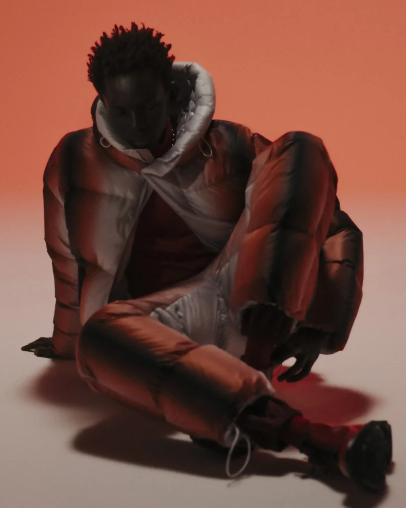 A model poses seated on the floor against a soft orange backdrop, wearing a gradient puffer jacket and pants in copper and silver tones from TATRAS’ “Layers of Japanese Craft” collection. The lighting accentuates the reflective textures and sculptural layering that define the capsule’s artisanal design