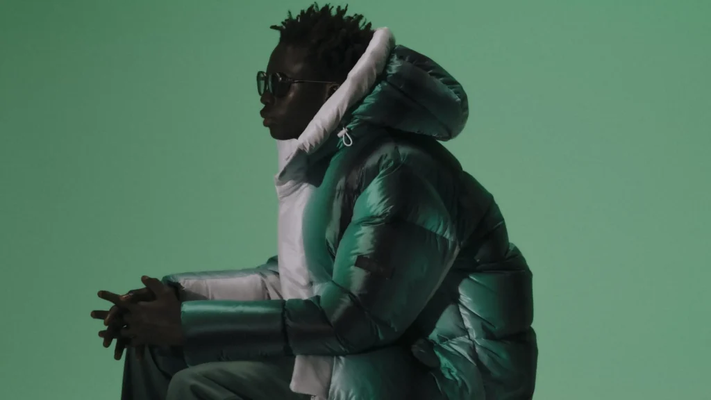 A model sits in profile against a muted green background, wearing a glossy, deep emerald puffer jacket with a high white hood from TATRAS’ “Layers of Japanese Craft” collection. The soft lighting and monochromatic tone emphasize the jacket’s sculptural silhouette and refined Japanese craftsmanship