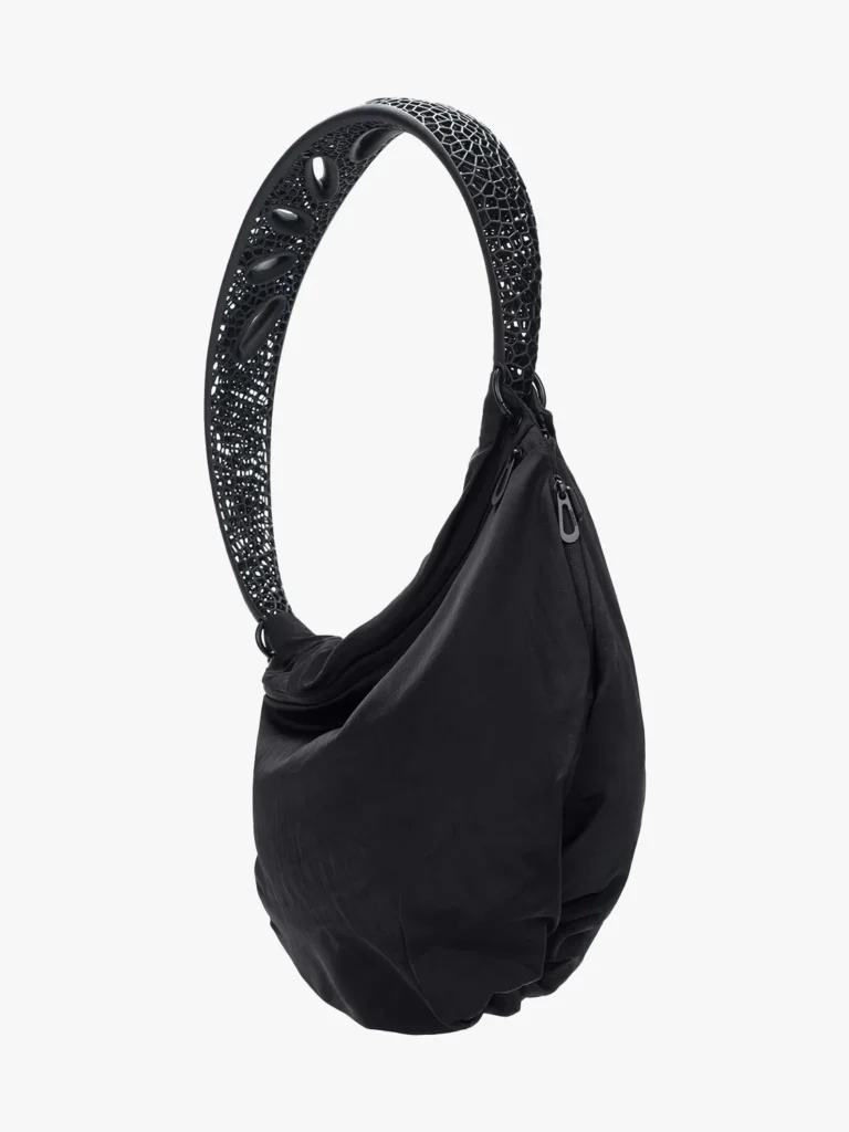 the nebula sling bag from the aliveform × côte&ciel “living systems” capsule, featuring a sculptural black nylon body contrasted by a wide 3d-printed lattice strap with organic perforations that curve like a spine, merging soft fabric with futuristic structural design