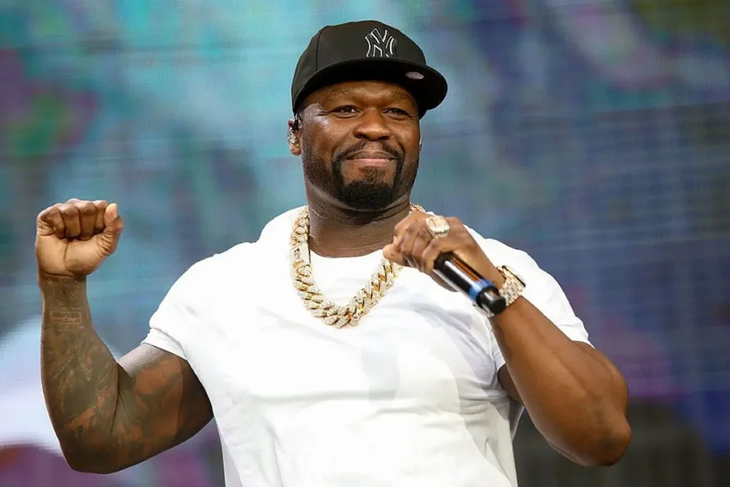 50 Cent performing on stage in a white tee and black cap, wearing a gold chain, captured mid-gesture with a confident smile — embodying the energy and charisma behind his new single Fightland with Sheff G, Sleepy Hallow, and Jeremih