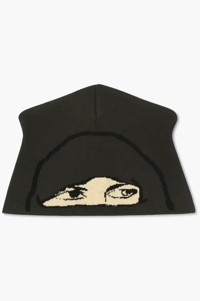 Black knit skully beanie displayed on a white background, featuring a beige woven graphic of a hooded figure’s eyes across the front