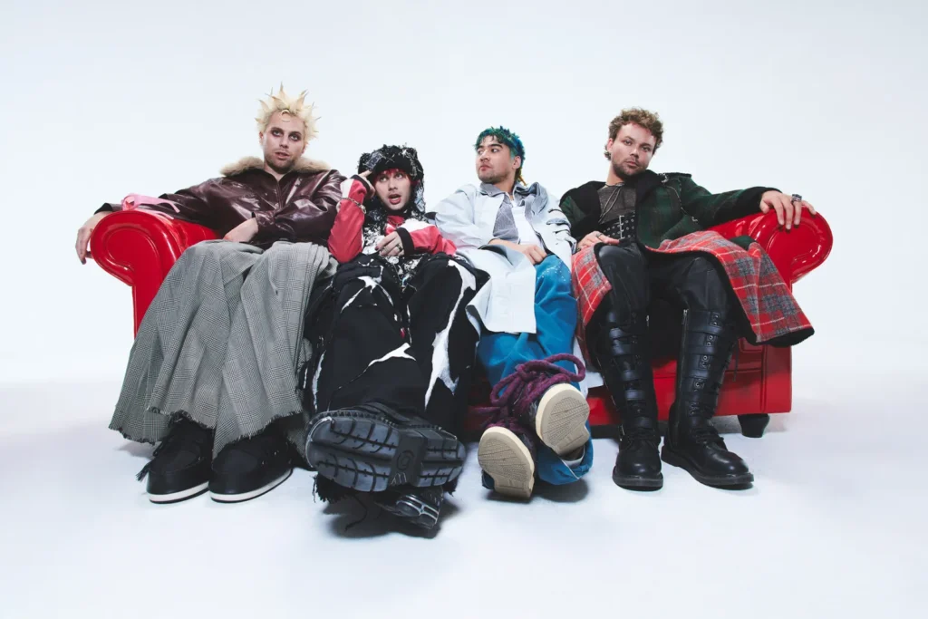 5 Seconds of Summer sitting on a glossy red couch against a white background, each member dressed in eclectic, avant-garde outfits with layered textures, bold patterns, and oversized silhouettes, looking relaxed and expressive