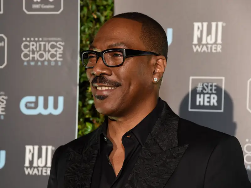 Eddie Murphy smiling on the red carpet at a Critics Choice Awards event, wearing black-framed glasses, a black patterned suit jacket, and a dark shirt, with sponsor logos and a leafy backdrop behind him