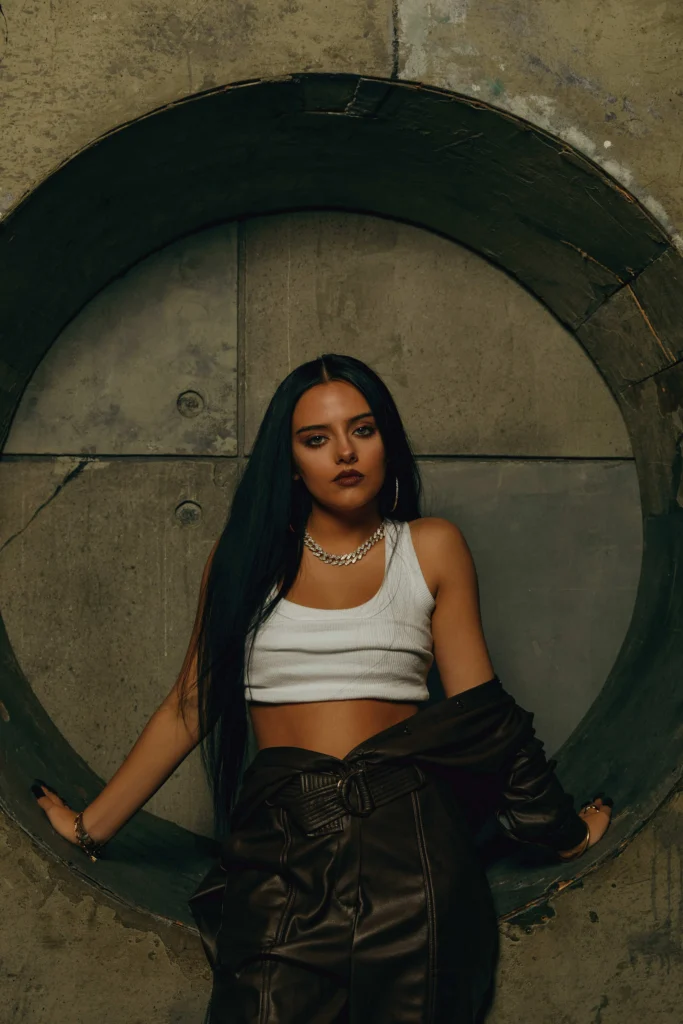 Jessie Murph with long dark hair stands confidently inside a large circular concrete frame, wearing a white crop tank top, dark leather pants, and layered jewelry, set against an industrial textured wall