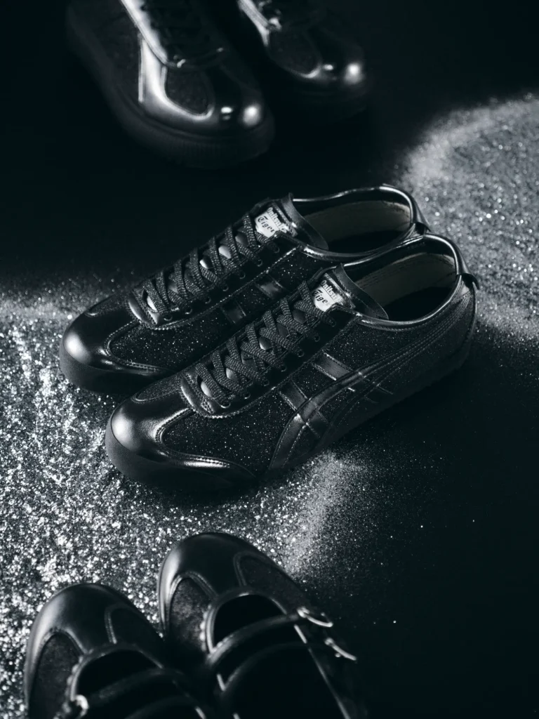 A pair of black Onitsuka Tiger sneakers with a subtle glitter finish rests on a dark surface lightly dusted with sparkling particles. The shoes feature glossy leather panels, tonal black Tiger Stripes, and a shimmering texture that catches the light. Additional black pairs appear blurred in the background, creating a moody, refined studio atmosphere