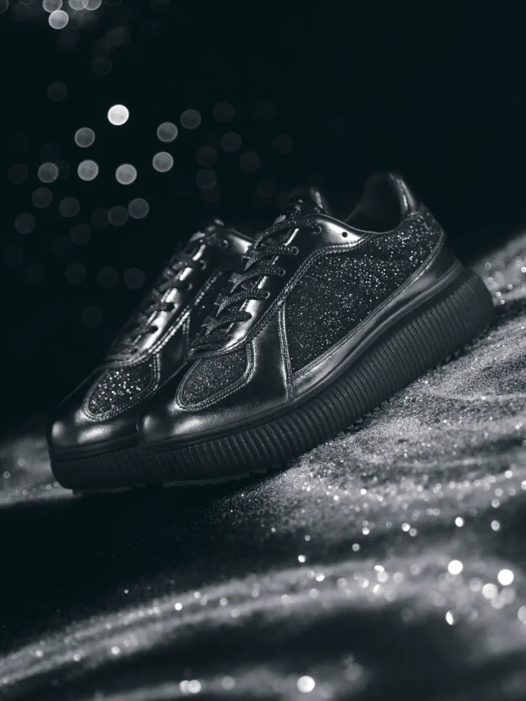 A pair of black Onitsuka Tiger sneakers with glitter-coated side panels and suede-like glitter overlays sits on a shimmering textured surface. The chunky black sole adds weight to the silhouette, while soft studio lighting creates sparkling reflections. Blurred bokeh lights glow in the dark background, enhancing the shoe’s glossy, night-inspired aesthetic