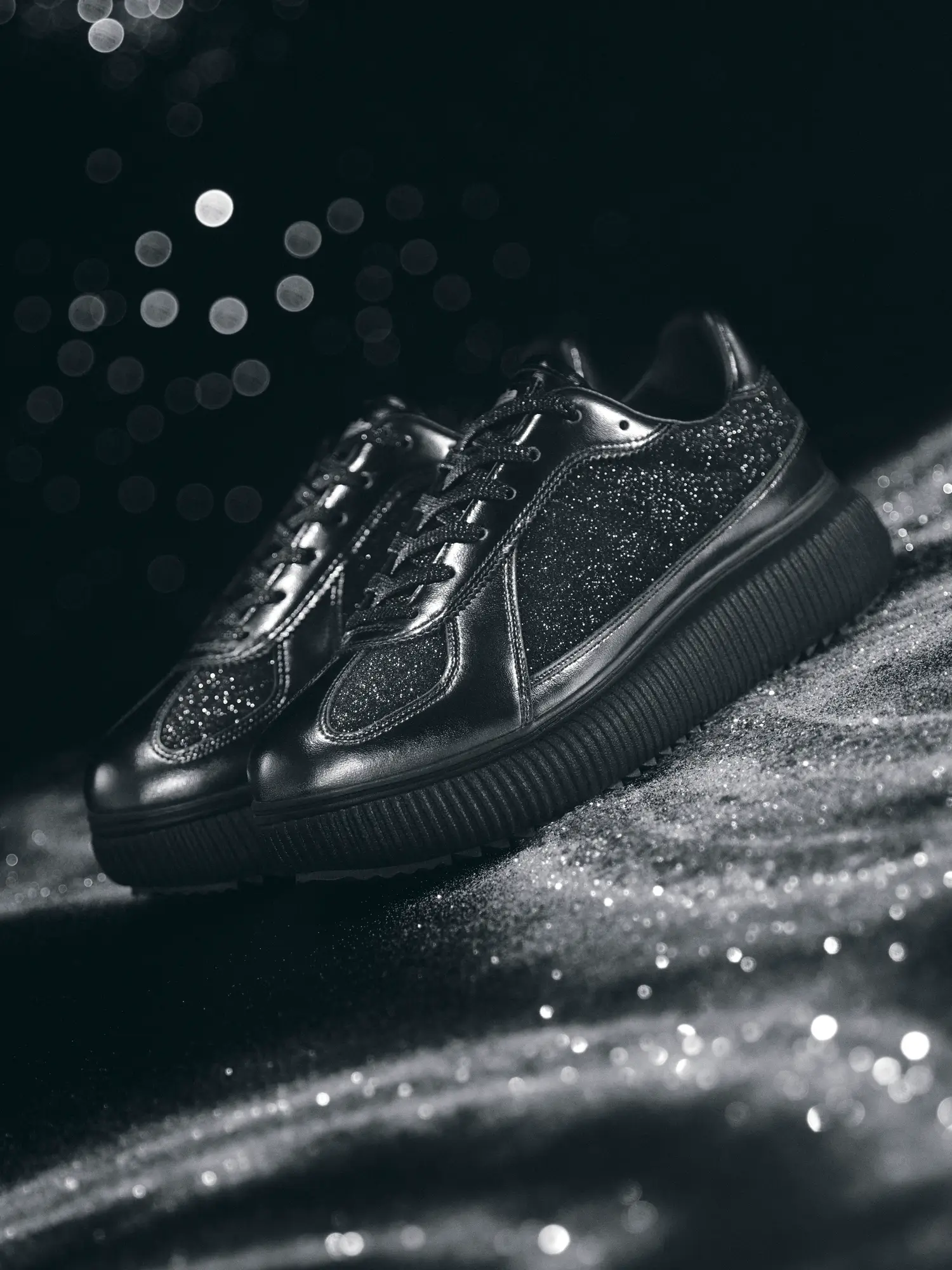 A pair of black Onitsuka Tiger sneakers with glitter-coated side panels and suede-like glitter overlays sits on a shimmering textured surface. The chunky black sole adds weight to the silhouette, while soft studio lighting creates sparkling reflections. Blurred bokeh lights glow in the dark background, enhancing the shoe’s glossy, night-inspired aesthetic
