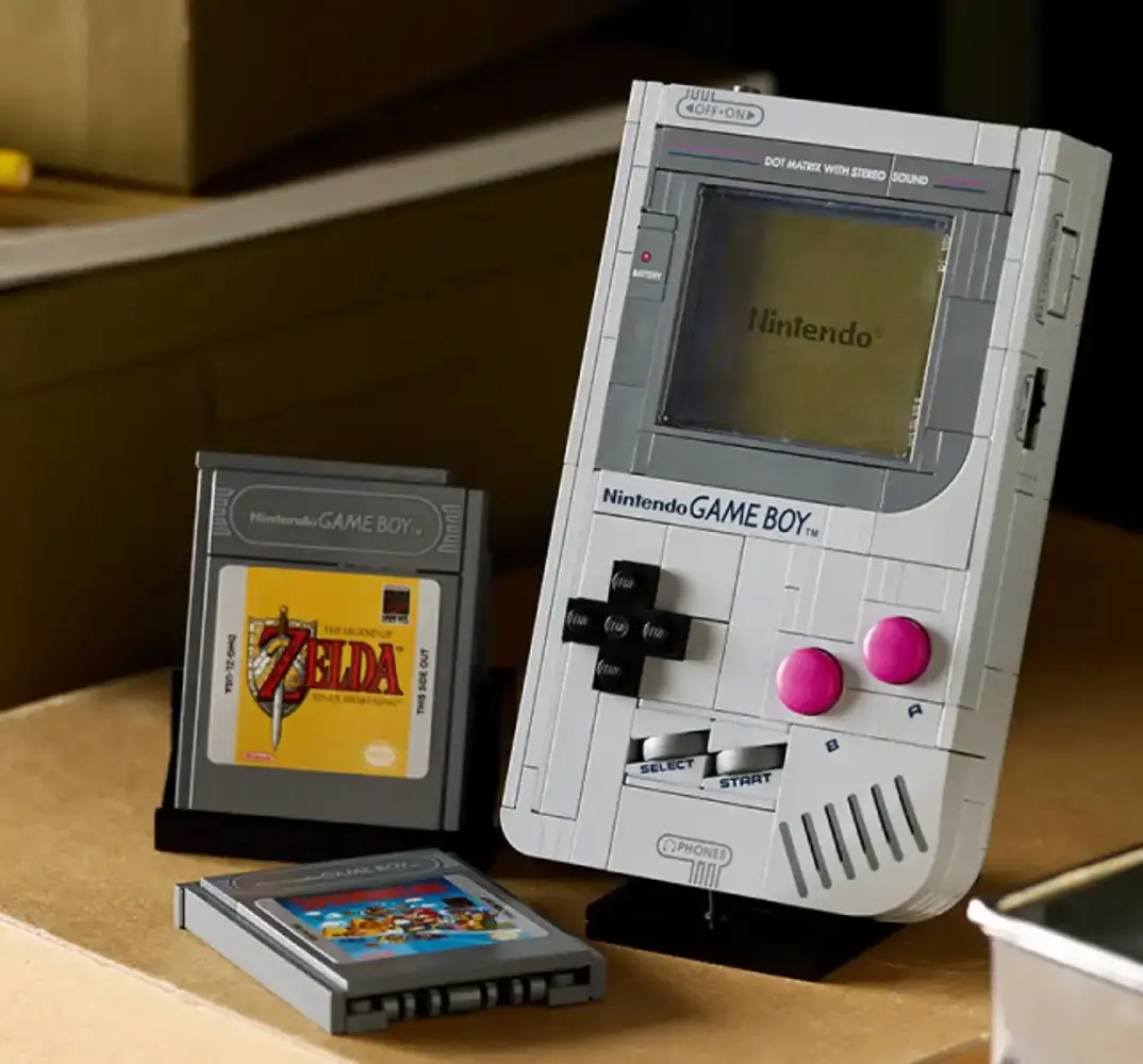 A LEGO-built replica of the classic Nintendo Game Boy stands on a black display base beside brick-built cartridges for The Legend of Zelda: Link’s Awakening and Super Mario Land, arranged on a wooden surface in warm ambient lighting