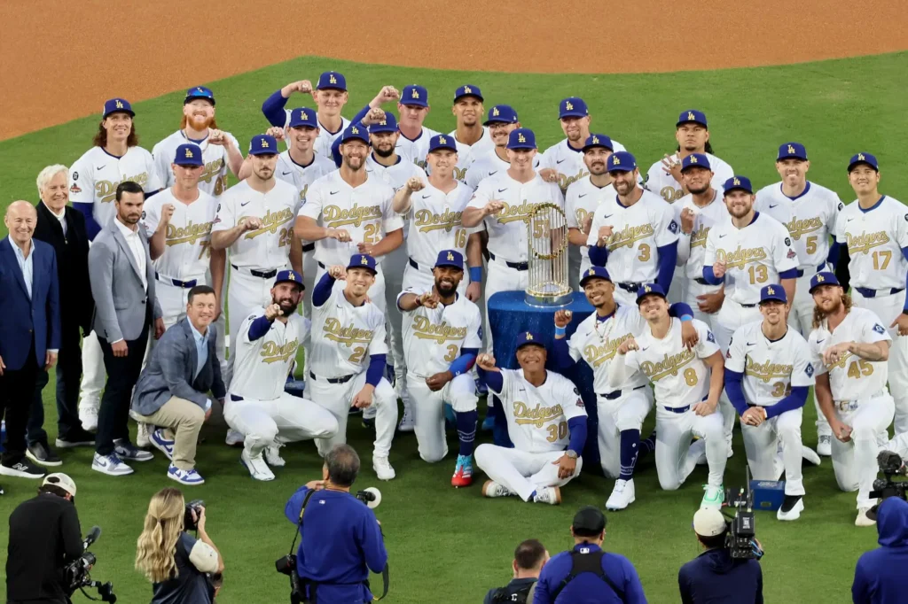 The Los Angeles Dodgers team poses together on the field with the 2025 World Series trophy, wearing white uniforms and blue caps, celebrating their championship victory while photographers capture the moment
