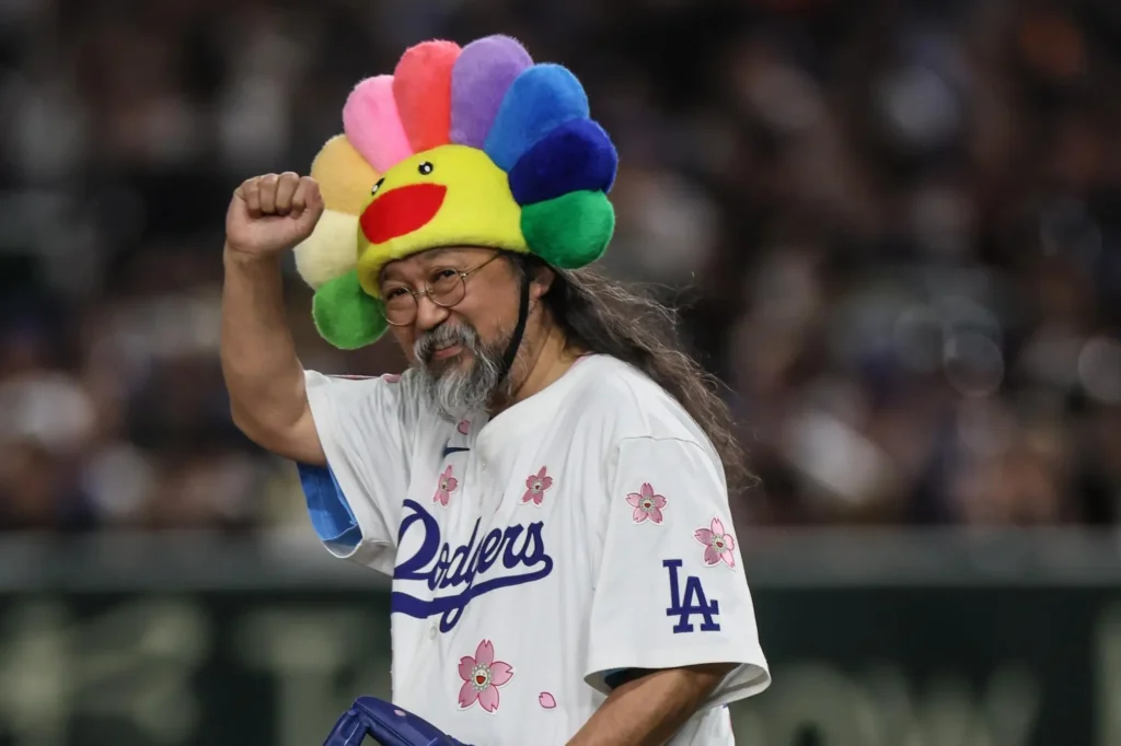Takashi Murakami wears a Los Angeles Dodgers jersey decorated with pink cherry blossoms and a plush rainbow flower hat, raising his fist and smiling during a celebratory appearance at the stadium