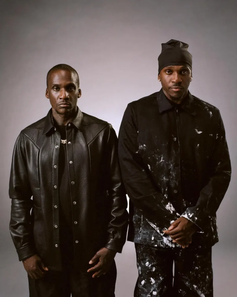 Clipse duo Pusha T and Malice pose against a neutral studio backdrop wearing black textured outfits, combining leather and paint-splattered fabrics in a minimalist portrait style