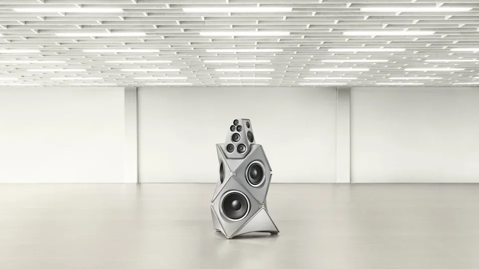A single Bang & Olufsen Beolab 90 Titan Edition speaker stands centered in a vast, minimalist gallery-like room with a geometric ceiling and soft neutral lighting, emphasizing its futuristic titanium form and multi-driver design