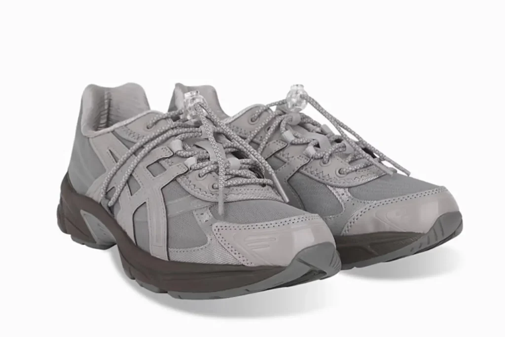A pair of ASICS GEL-1130 RE sneakers in a monochromatic grey colorway, featuring layered mesh and synthetic panels, rounded toe guards, reflective detailing, and thick patterned laces secured with translucent toggle locks. The shoes sit on a dark grey midsole with a sculpted outsole, photographed against a clean white background