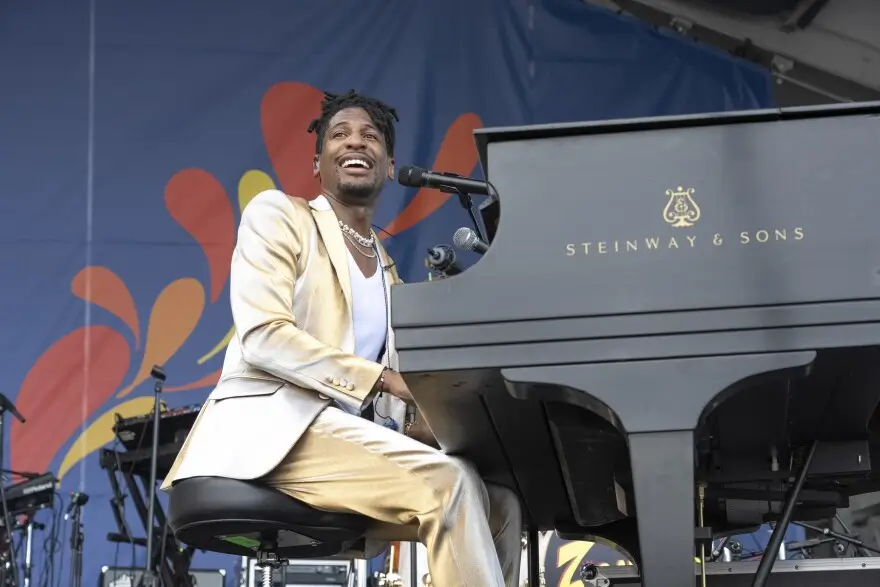 Jon Batiste performs onstage wearing a gold suit, smiling as he plays a black Steinway & Sons grand piano against a colorful festival backdrop