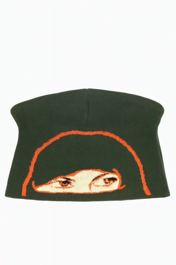 Olive-green knit skully beanie displayed against a white background, featuring an orange graphic of a hooded figure’s eyes woven across the front