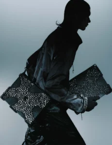 a model dressed in glossy black fabric carries two pieces from the aliveform × côte&ciel “living systems” capsule. the accessories feature intricate black 3d-printed lattice shells that resemble cellular structures, blending organic patterning with engineered precision against a muted gradient background