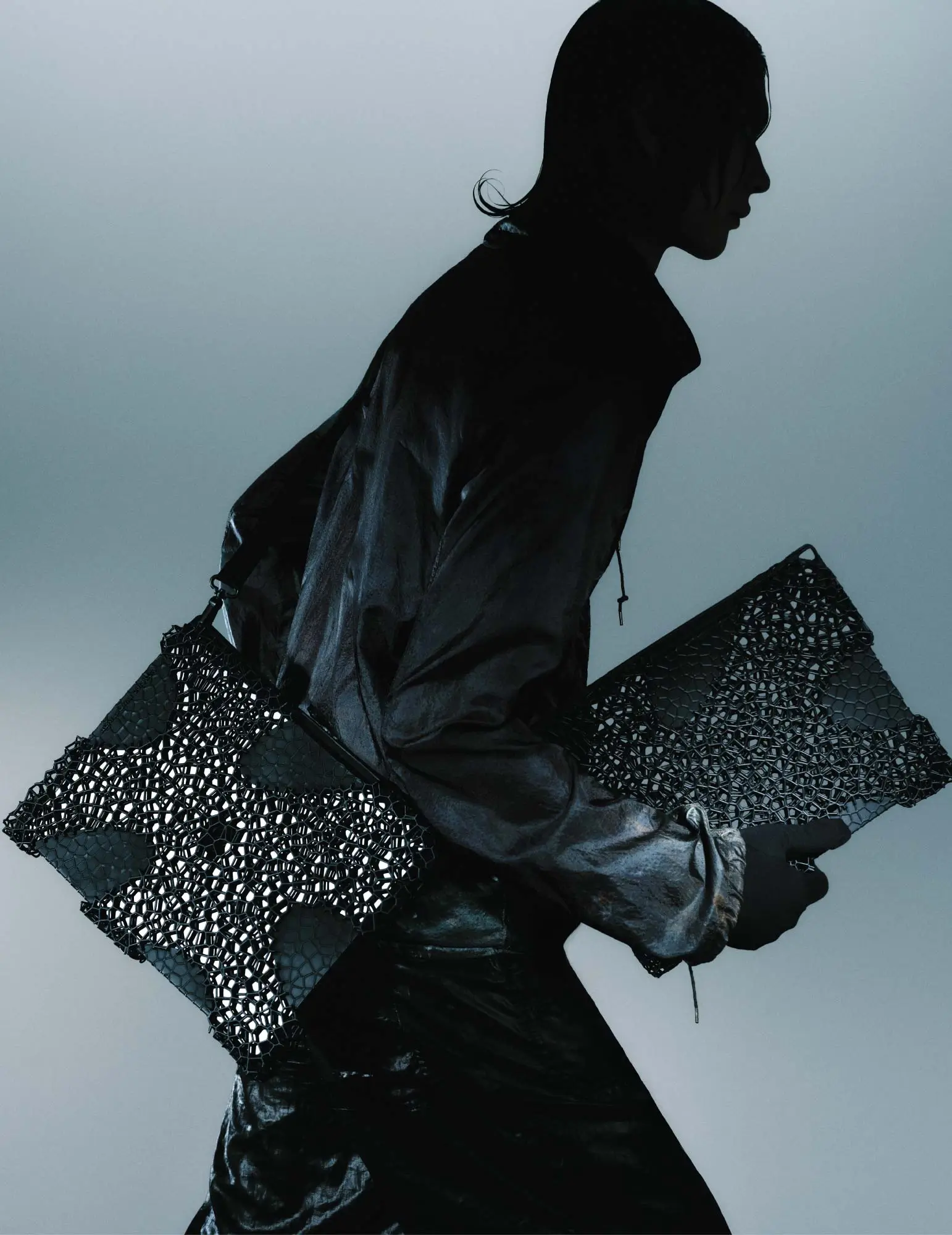 a model dressed in glossy black fabric carries two pieces from the aliveform × côte&ciel “living systems” capsule. the accessories feature intricate black 3d-printed lattice shells that resemble cellular structures, blending organic patterning with engineered precision against a muted gradient background