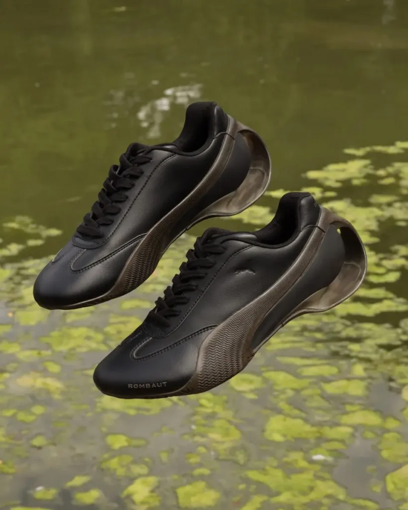 Black PUMA x ROMBAUT Season 2 sneakers floating above a green, algae-covered pond, highlighting the shoe’s distressed brown tooling, smooth synthetic leather upper, and sculptural cell-inspired heel design