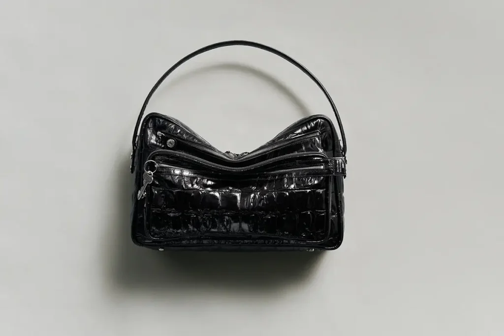 Acne Studios FW25 Holiday Gift Selection — a glossy black crocodile-embossed leather shoulder bag with silver zip detailing and structured silhouette, photographed against a neutral grey background to highlight its sculptural form and minimalist elegance