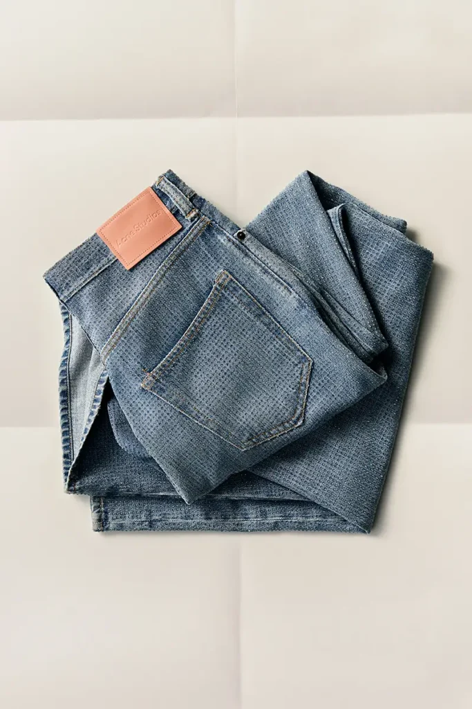 Acne Studios FW25 Holiday Gift Selection — a pair of light blue textured denim jeans neatly folded on a neutral backdrop, featuring the signature pink Acne Studios leather patch, symbolizing the brand’s clean and enduring Scandinavian craftsmanship