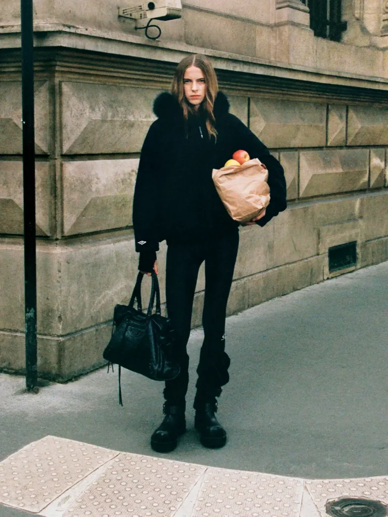 Model wearing an all-black Balenciaga Skiwear 2025 outfit with fur-trimmed hood, holding a grocery bag of apples and carrying a black Le City bag, photographed on a Paris street for Balenciaga’s Winter 25 ski campaign by Aidan Zamiri