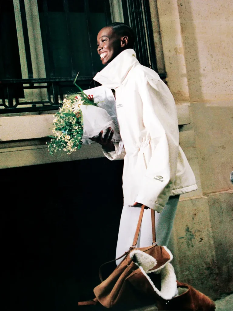 Model wearing an off-white Balenciaga Skiwear 2025 jacket while smiling and holding a bouquet of flowers on a Paris street, carrying a shearling-lined brown duffle bag; photographed by Aidan Zamiri for Balenciaga’s Winter 25 ski campaign