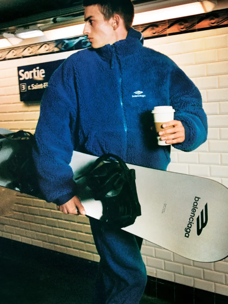 Model wearing an off-white Balenciaga Skiwear 2025 jacket while smiling and holding a bouquet of flowers on a Paris street, carrying a shearling-lined brown duffle bag; photographed by Aidan Zamiri for Balenciaga’s Winter 25 ski campaign