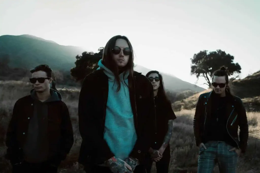 The band Bad Omens standing outdoors in a muted, cinematic landscape with mountains and dry grass behind them. All four members wear dark clothing and sunglasses, with the lead figure centered in a light gray hoodie and jacket. The evening light casts cool tones across the scene, giving the group a moody, atmospheric presence