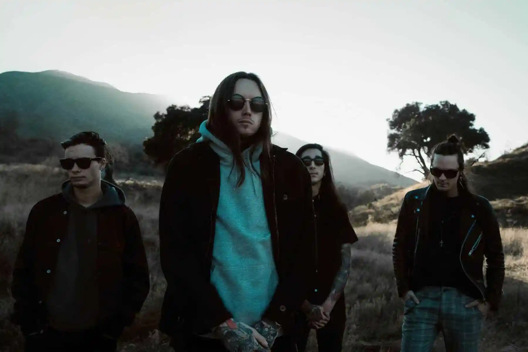 The band Bad Omens standing outdoors in a muted, cinematic landscape with mountains and dry grass behind them. All four members wear dark clothing and sunglasses, with the lead figure centered in a light gray hoodie and jacket. The evening light casts cool tones across the scene, giving the group a moody, atmospheric presence