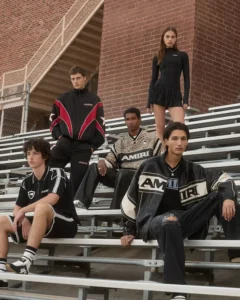 A group of five young models sit and stand on metal bleachers outside a brick school building, dressed in AMIRI Pre-Spring 2026 pieces including varsity knits, athletic sets, track jackets, and a leather logo jacket, styled in a cinematic, youth-culture aesthetic