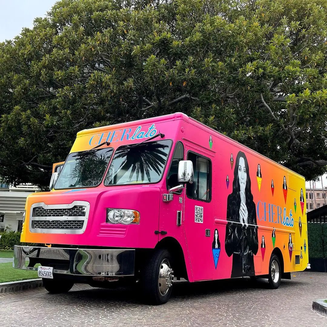 CherLato gelato truck parked under a large tree in Los Angeles, featuring a bright pink-to-orange gradient wrap with Cher’s black-and-white portrait and colorful diamond motifs, representing her artisan gelato brand