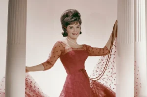 Connie Francis posing elegantly between two white columns, wearing a red polka-dot tulle dress with pearl necklace, captured in a 1960s studio portrait that embodies the soft glamour of her “Pretty Little Baby” era