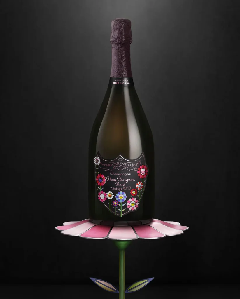A Dom Pérignon Rosé Vintage 2010 champagne bottle from the Takashi Murakami limited edition rests atop a sculptural pink flower pedestal. The dark bottle features Murakami’s vibrant smiling flower motifs in shades of red, pink, and white, symbolizing the joy fusion of haute champagne artistry and contemporary Japanese pop culture.