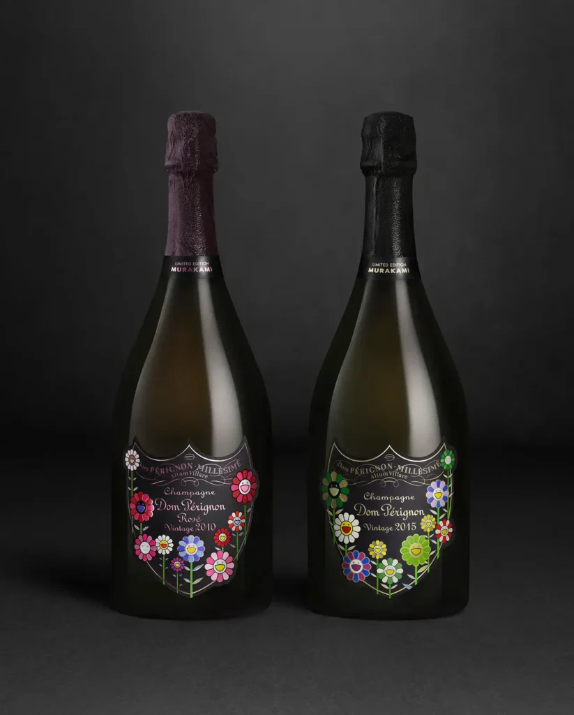 Two limited-edition Dom Pérignon x Takashi Murakami champagne bottles stand side by side against a dark background. The left bottle, Rosé Vintage 2010, features Murakami’s vibrant red and pink smiling flowers, while the right, Vintage 2015, showcases multicolored blooms. Both designs merge Dom Pérignon’s classic elegance with Murakami’s joy pop-art aesthetic.