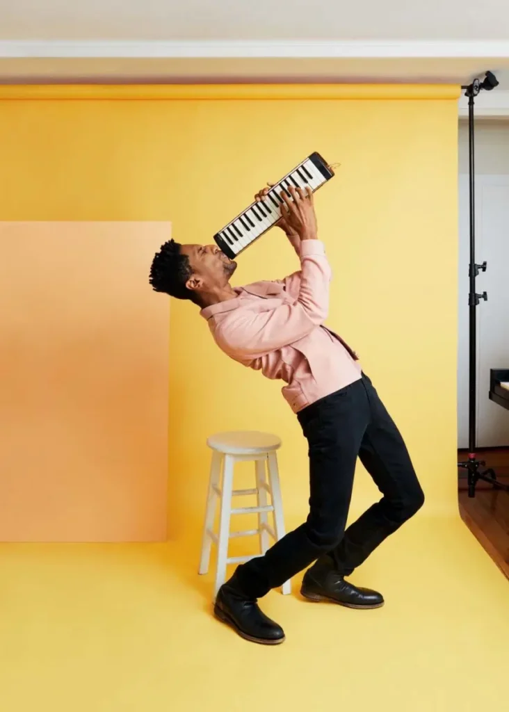 Jon Batiste leans back playfully while performing on a melodica in a studio with a bright yellow backdrop, dressed in a pink jacket and black jeans