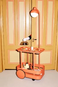 An orange Fatboy Jolly Trolley bar cart styled indoors with a built-in curved LED lamp, displaying breakfast items on the upper tray while a gloved hand opens a door behind it in a vintage mustard-and-peach room