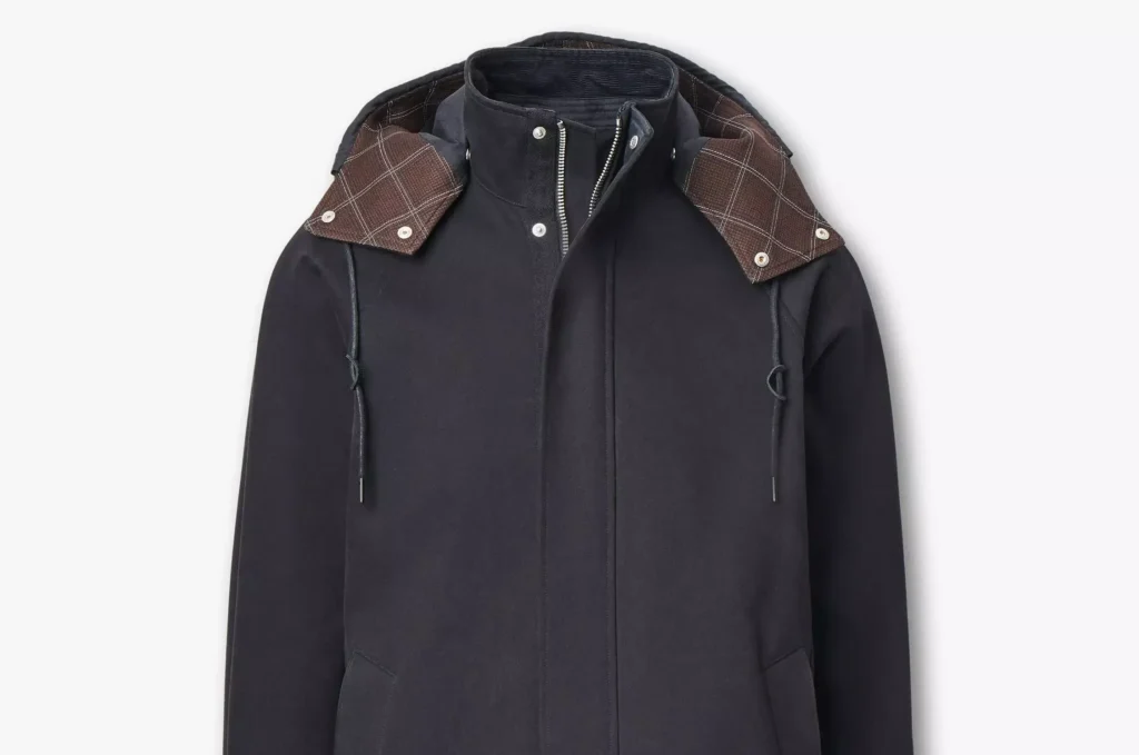 front close-up of the Rag & Bone Garret Coat in matte black, showing its structured hood lined with brown plaid fabric, concealed zip placket, and minimalist tailored design