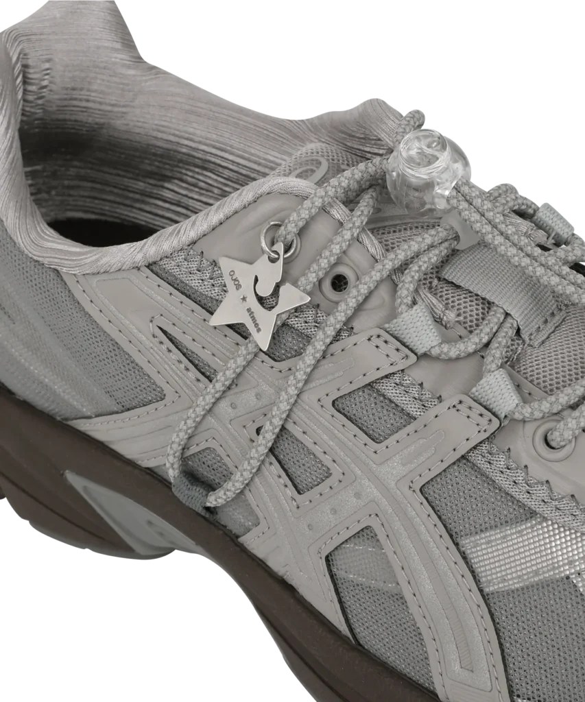 Close-up of the OJOS x atmos Pink x ASICS GEL-1130 RE in grey, showing the layered mesh and synthetic overlays, patterned laces with a clear toggle, and a metallic star-shaped charm engraved with “OJOS × atmos.” The textured collar and dark midsole are also visible, highlighting the shoe’s detailed construction