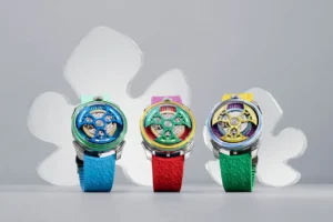A trio of Yinka Ilori × MAD Editions × MB&F watches displayed upright against a soft grey backdrop, each featuring vibrant multicolored open-worked dials and textured rubber straps in blue, red, and green. The watches showcase Ilori’s signature joyful palette, with bold geometric accents and transparent mechanics visible beneath domed sapphire crystals