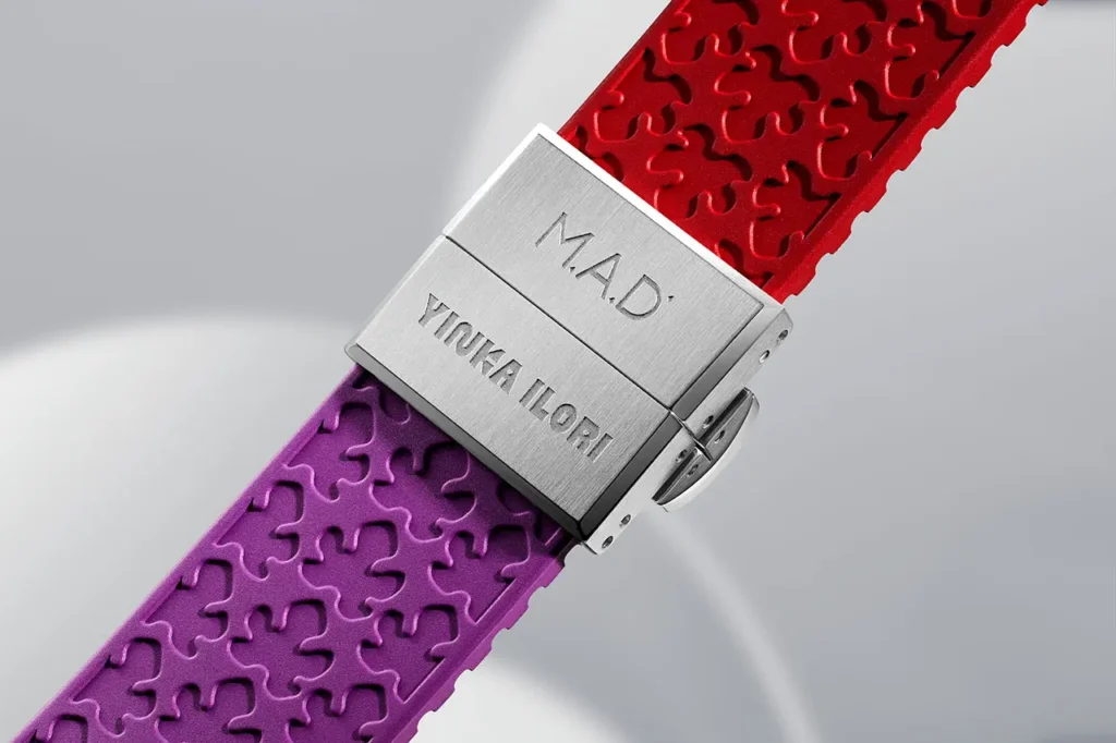 Close-up of the brushed metal clasp on the Yinka Ilori × MAD Editions × MB&F watch, engraved with “M.A.D.” and “YINKA ILORI.” The clasp connects a purple and red textured rubber strap featuring Ilori’s signature repeating pattern, shown against a soft grey, curved backdrop