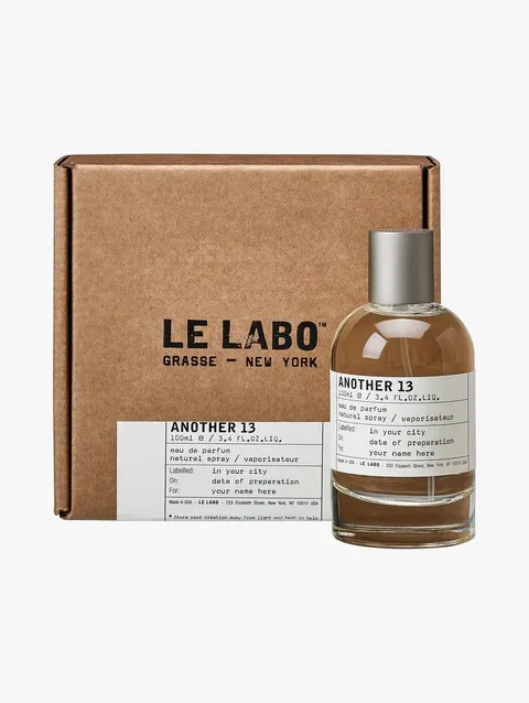 A clear glass bottle of Le Labo AnOther 13 Eau de Parfum with a silver metal cap and minimalist white label sits in front of its brown kraft-cardboard box. The box features bold black Le Labo branding and a white ingredient-style sticker, reflecting the brand’s industrial, lab-inspired aesthetic