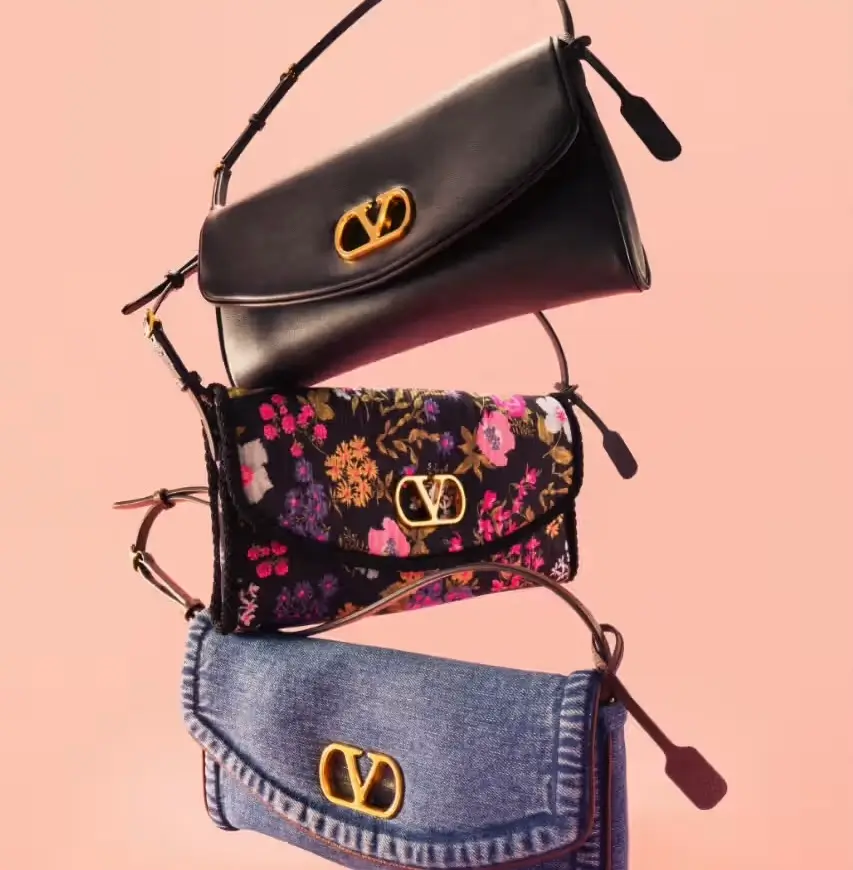 Three Valentino Garavani DeVain bags stacked vertically against a soft pink background, featuring a smooth black leather version on top, a floral embroidered style in the middle, and a blue denim edition at the bottom, each adorned with the signature gold VLogo buckle