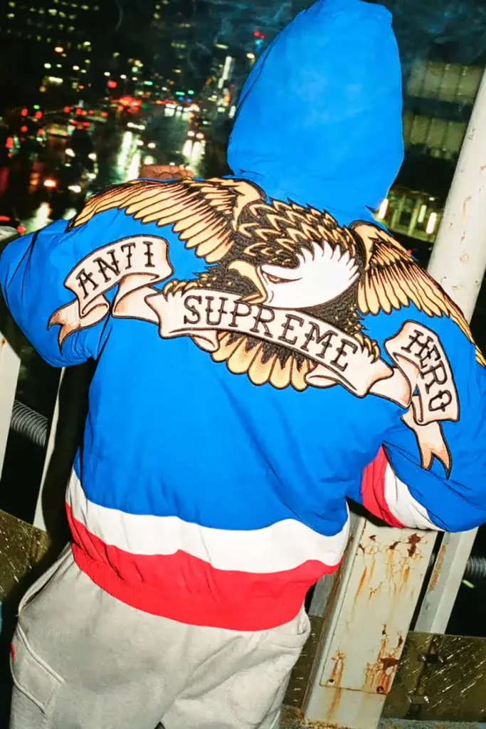 A person wearing a blue hooded jacket stands overlooking a rainy cityscape at night, with a large embroidered eagle graphic on the back featuring banners reading “ANTI,” “SUPREME,” and “HERO,” accented by red and white panels at the hem
