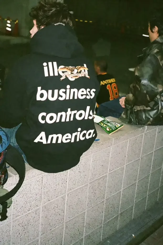 A person sits on a tiled ledge wearing a black Supreme × Antihero hoodie featuring the bold back print “ill business controls America.” with a large co-branded eagle graphic, while others in streetwear sit nearby in an urban nighttime setting