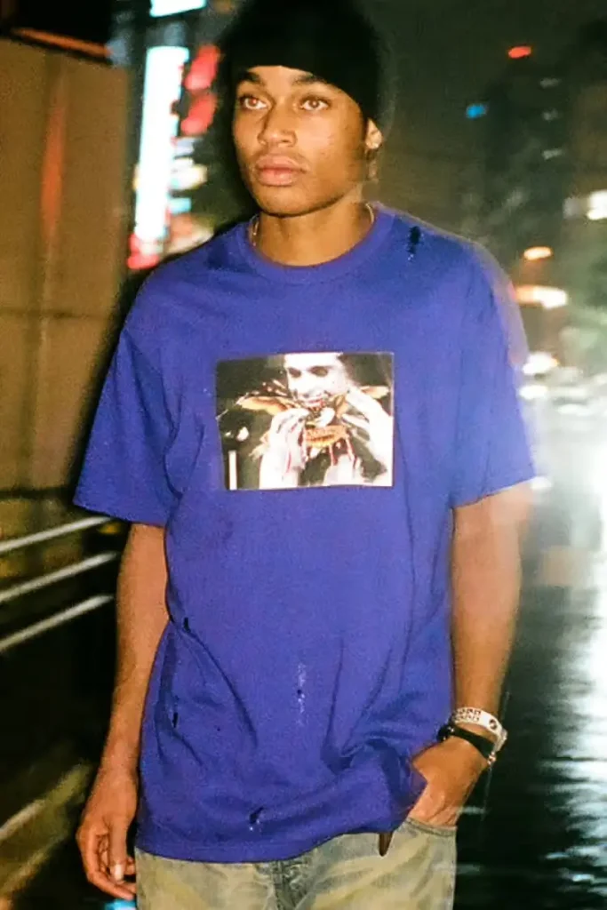 A person walks through a rainy city at night wearing a blue Supreme × Antihero T-shirt featuring a graphic print on the chest, paired with light denim and a black beanie, illuminated by blurred street lights in the background