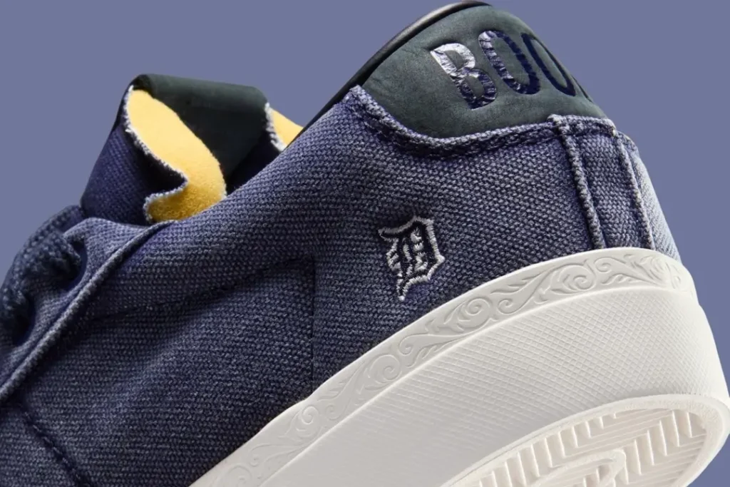 Close-up of a navy blue sneaker showing textured canvas material, an embroidered monogram-style logo near the heel, and a white midsole with subtle embossed detailing