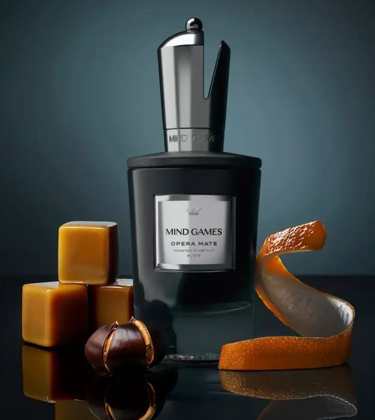 Mind Games Opera Mate perfume bottle displayed with caramel cubes, roasted chestnuts, and a spiral of orange peel on a reflective dark surface, symbolizing the fragrance’s warm and gourmand notes
