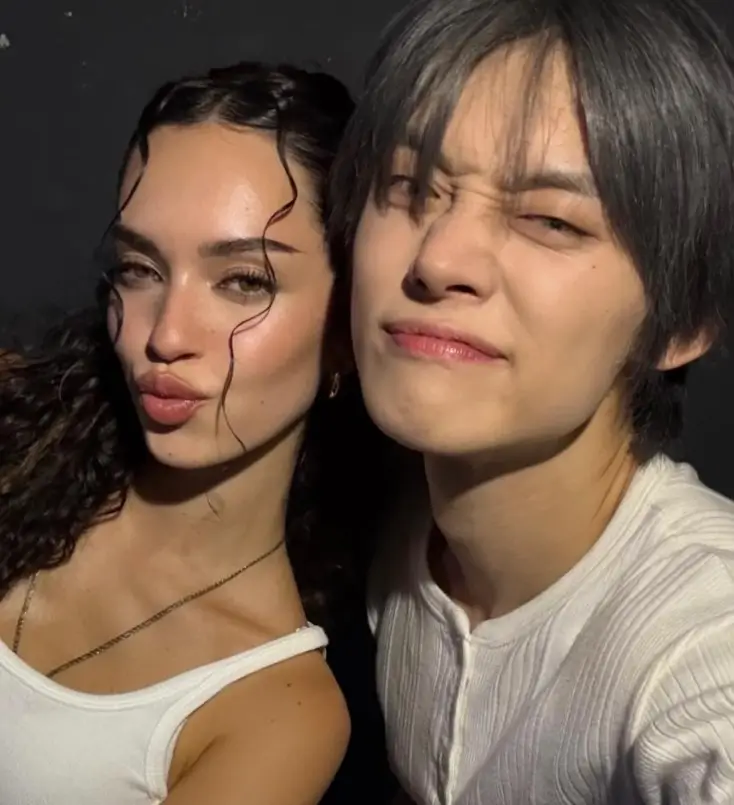 Yeonjun and Daniela pose together in a candid backstage photo, both wearing white tops under soft lighting. They lean close, smiling playfully — Yeonjun squints slightly while Daniela makes a pout, capturing a casual yet intimate energy that mirrors the chemistry of their lab Let Me Tell You