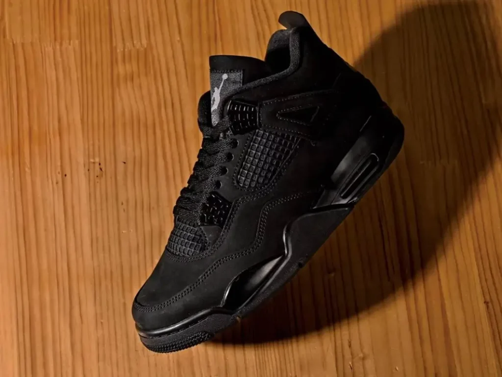 Air Jordan 4 “Black Cat” sneaker photographed on a wooden surface, showcasing its all-black nubuck upper, mesh panels, and tonal sculpted midsole in dramatic lighting