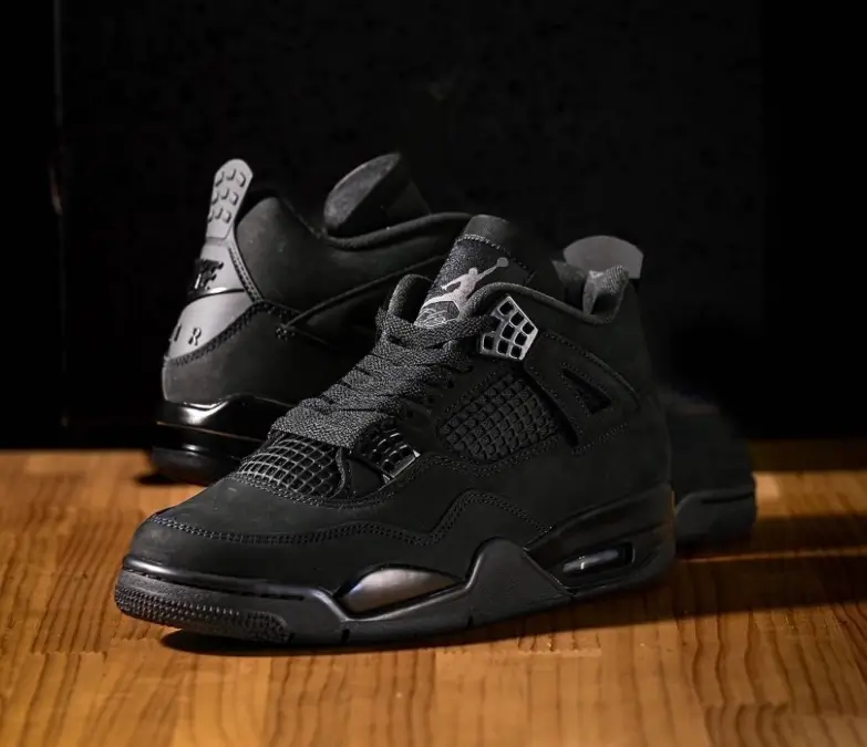 Air Jordan 4 “Black Cat” sneakers displayed on a wooden surface, featuring an all-black nubuck upper, black mesh panels, and tonal midsole with subtle metallic details